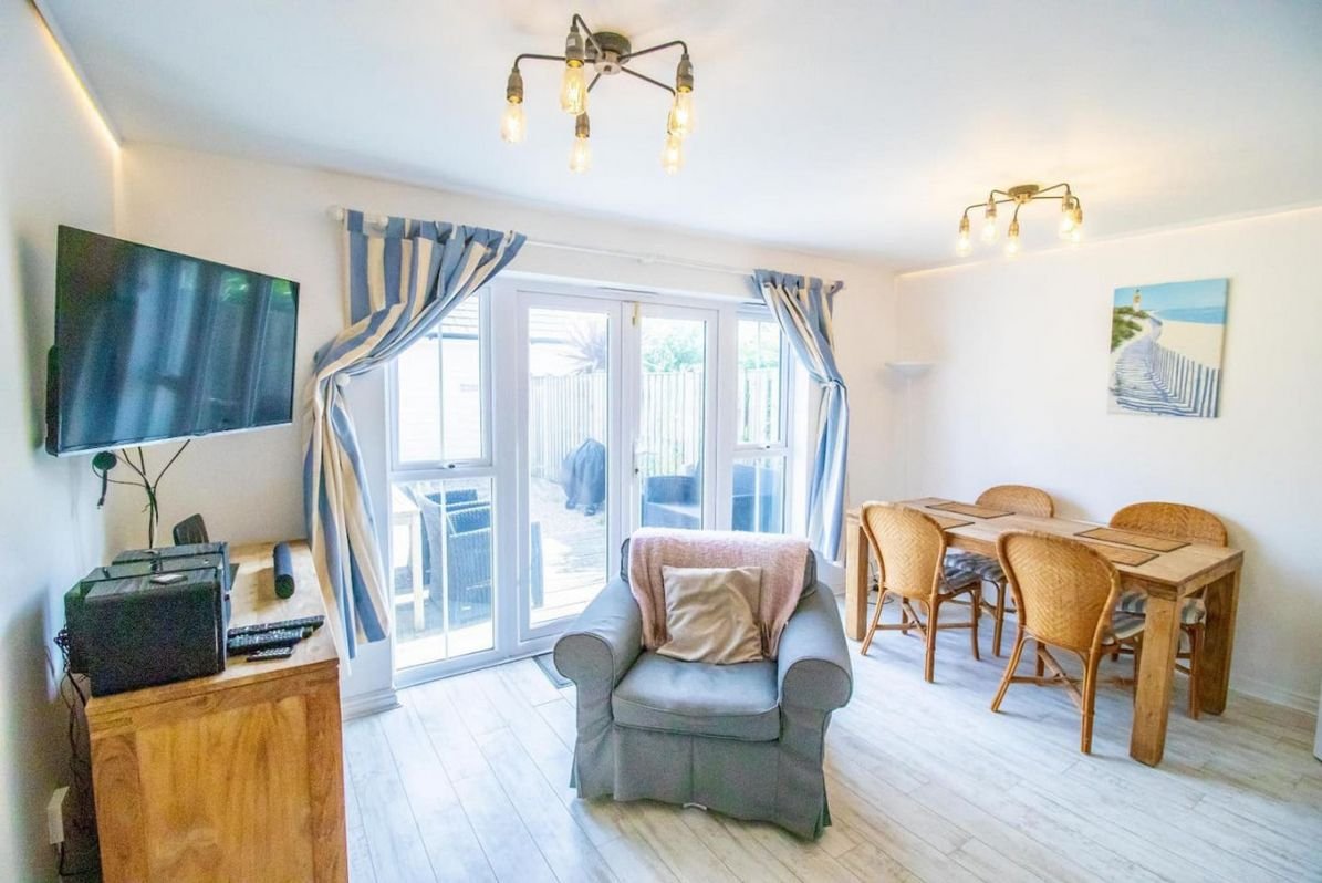 Sunlit lounge at Beachside Cottage with a grey armchair, dining table and French windows opening onto the decking area